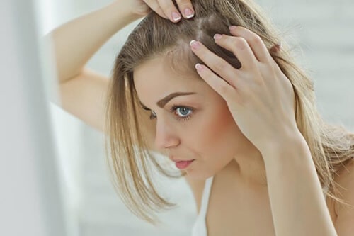 Women in the mirror parting hair to a bald spot and showing concern for hair loss treatments